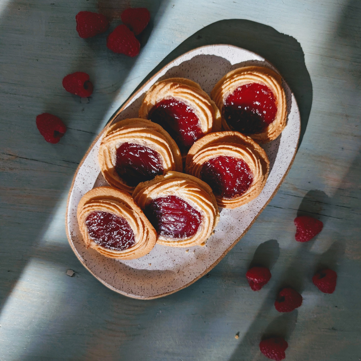 Six jam-filled butter cookies on an oval speckled plate, scattered raspberries on a sunlit blue wooden table