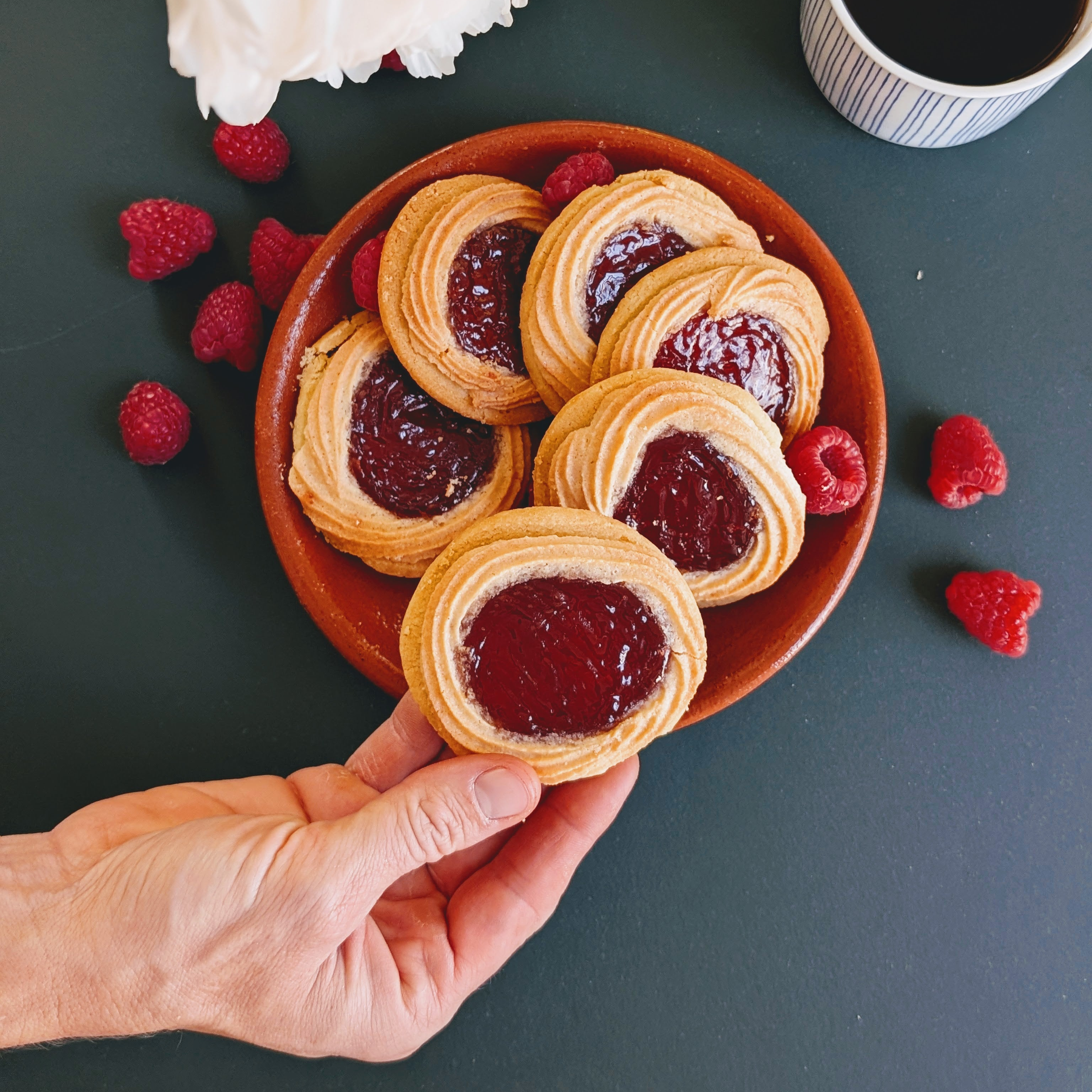 Hand holding a raspberry-jam thumbprint cookie above a plate of cookies, with scattered raspberries and a cup of coffee.