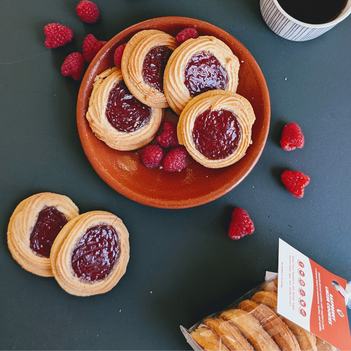 Jam-topped thumbprint cookies on a terracotta plate with fresh raspberries, scattered berries, a cup of coffee, and packaged cookies.