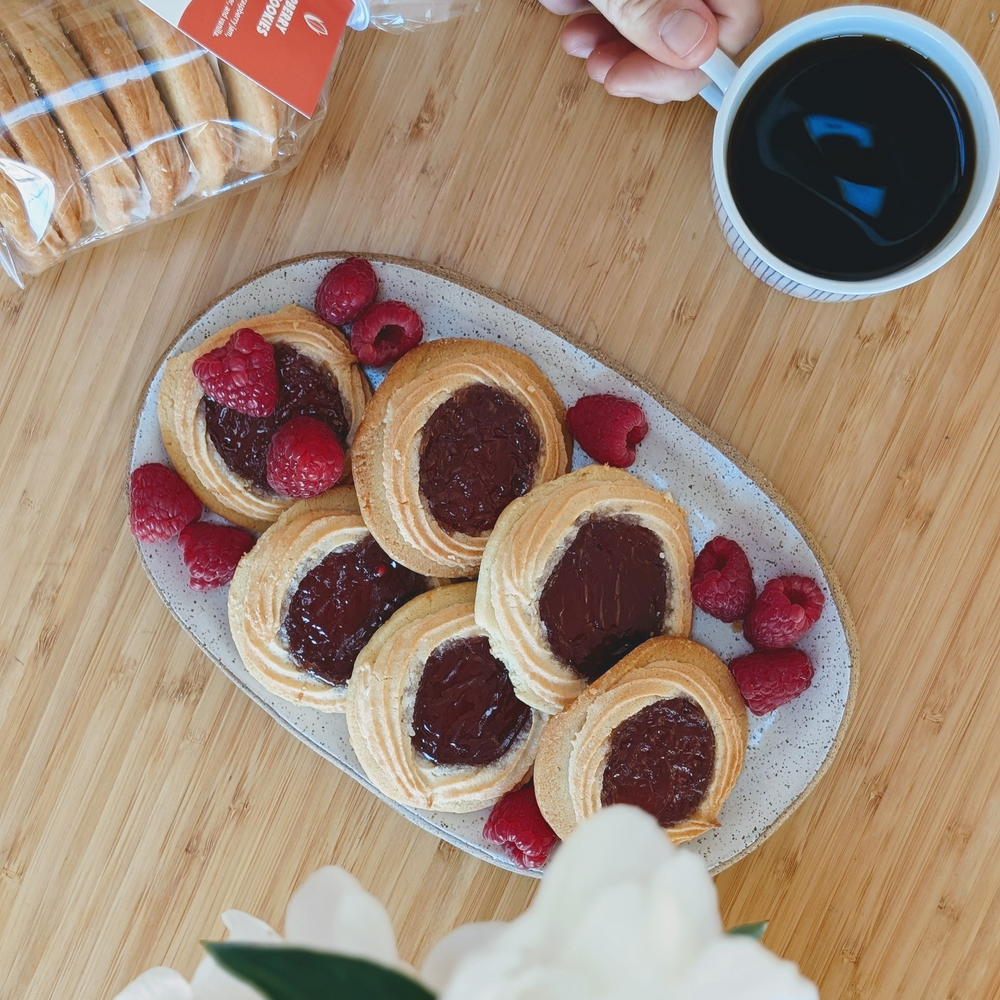 Plate of six jam-filled thumbprint cookies with raspberries and a cup of black coffee on a wooden table.