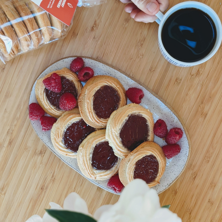 Plate of six jam-filled thumbprint cookies with raspberries and a cup of black coffee on a wooden table.