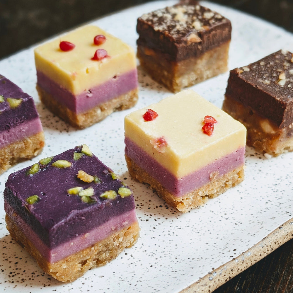 Layered dessert squares on a plate: crumb crust, purple layer, yellow topping with pomegranate seeds; chocolate squares behind.