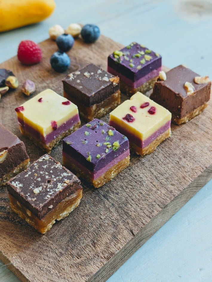 Colorful layered desserts on a wooden board, featuring chocolate, raspberry, and pistachio layers.