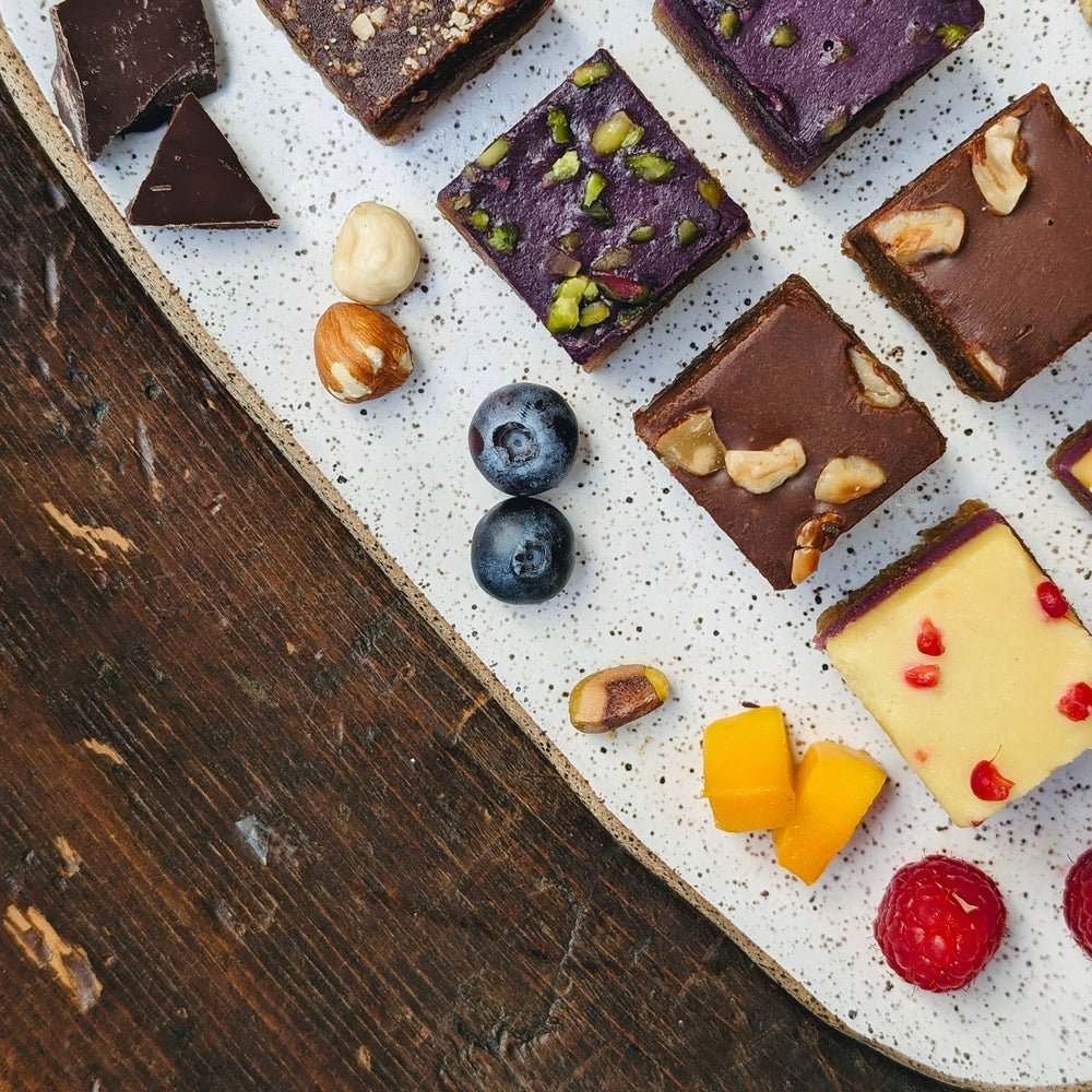 Assorted bite-size dessert squares, blueberries, raspberries, nuts and mango on a speckled white platter atop dark wood.