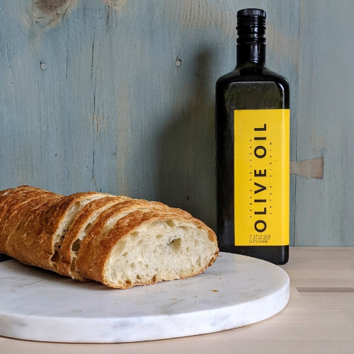 Sliced baguette on a marble board beside a dark bottle labeled OLIVE OIL.