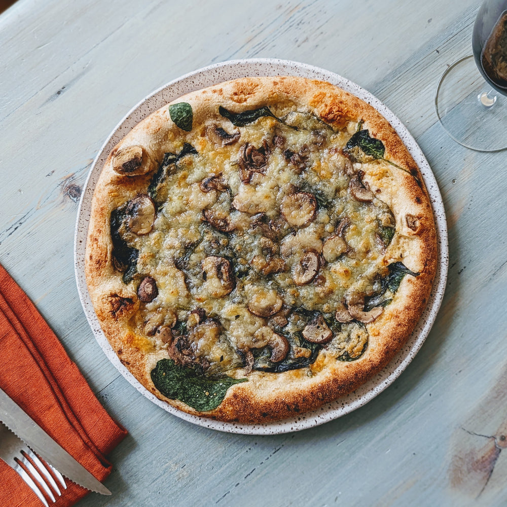 Mushroom and spinach pizza with melted cheese on a ceramic plate, napkin and fork at left and a wine glass at right