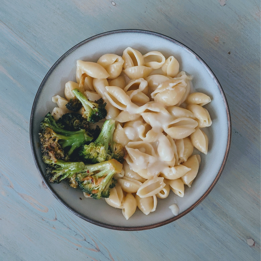 Top-down view of a bowl of shell pasta in creamy sauce with roasted broccoli on a light blue wooden table.