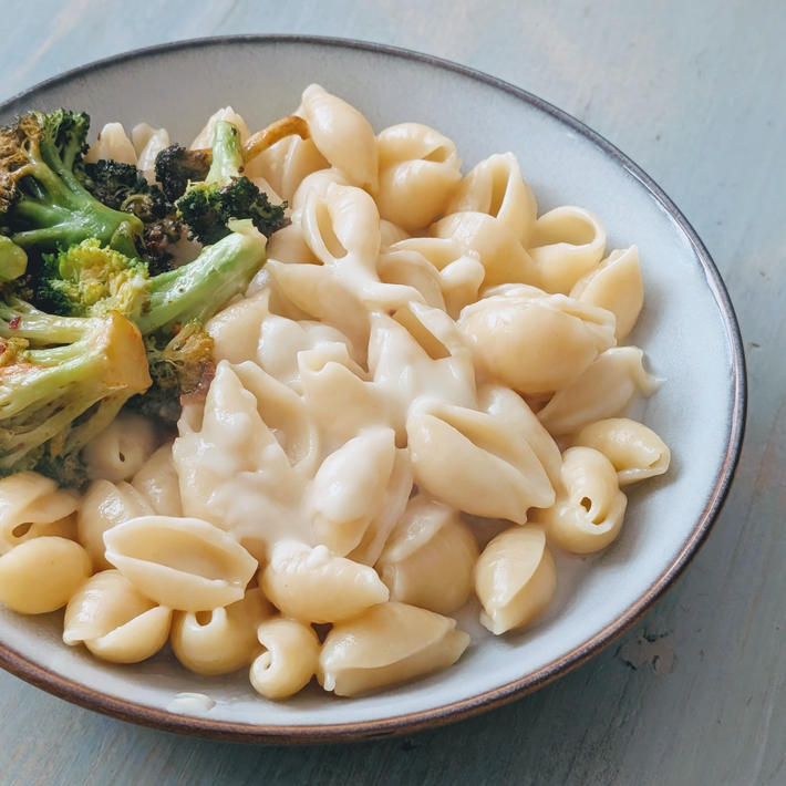 Overhead view of shell pasta coated in Wildgrain artisan three-cheese sauce with roasted broccoli in a blue-rimmed bowl on a pale wooden table