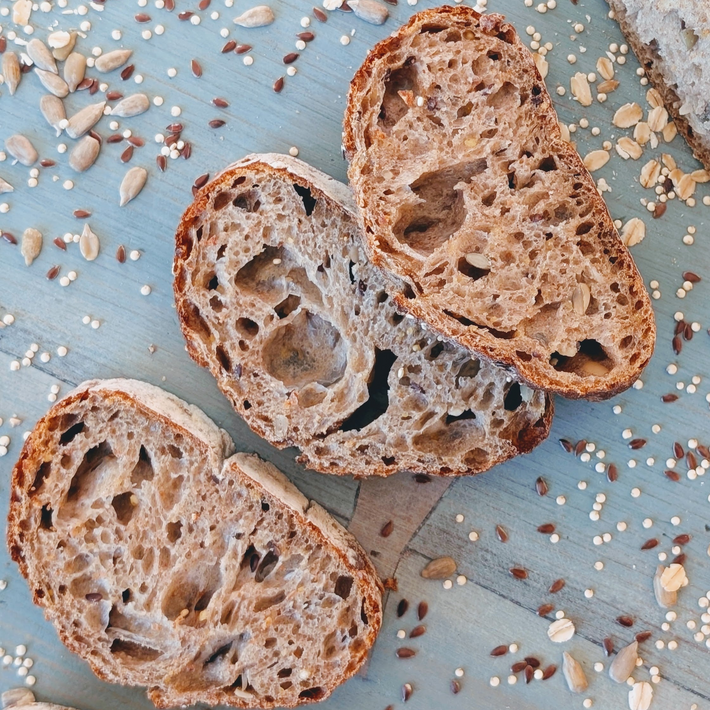 Three slices of seeded multigrain bread on a blue wooden surface with scattered sunflower seeds, flax, and oats