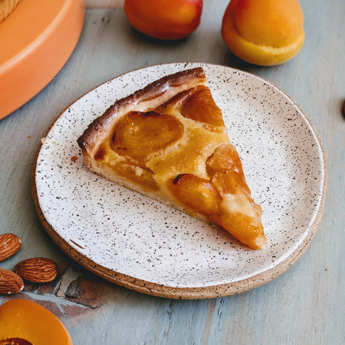Slice of apricot tart on a speckled ceramic plate, with fresh apricots and almonds on a blue wooden table.