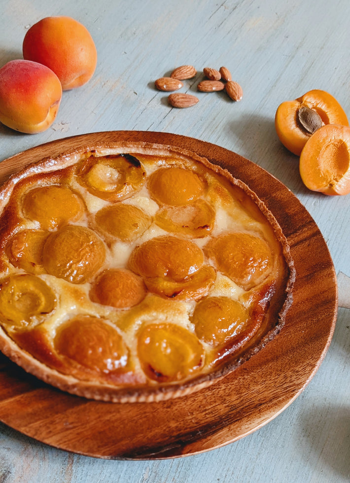 Apricot tart topped with halved glazed apricots on a wooden board, with whole apricots and almond kernels nearby