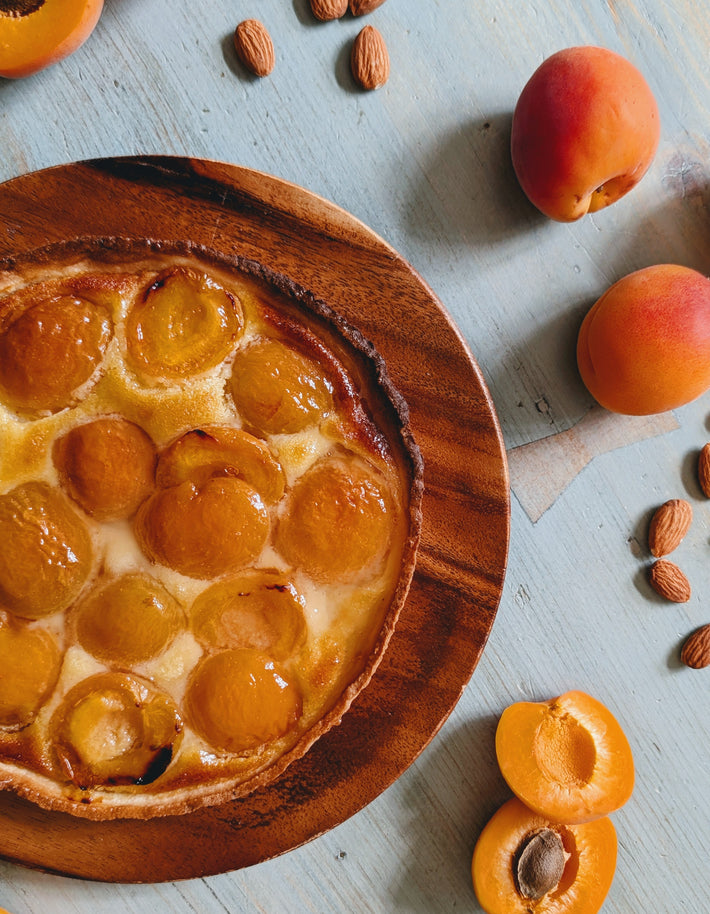 Apricot tart with glazed halved apricots on a round wooden plate, surrounded by whole apricots and almonds on a blue table.