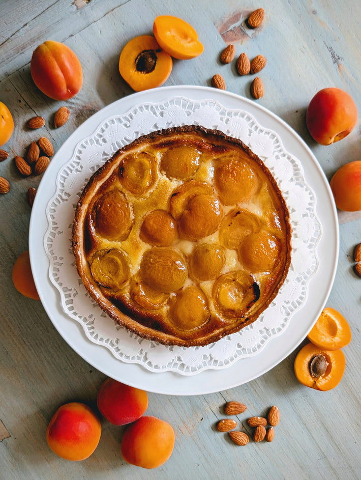 Apricot tart on a white plate with a paper doily, surrounded by whole apricots and almonds on a light wood table, top-down.