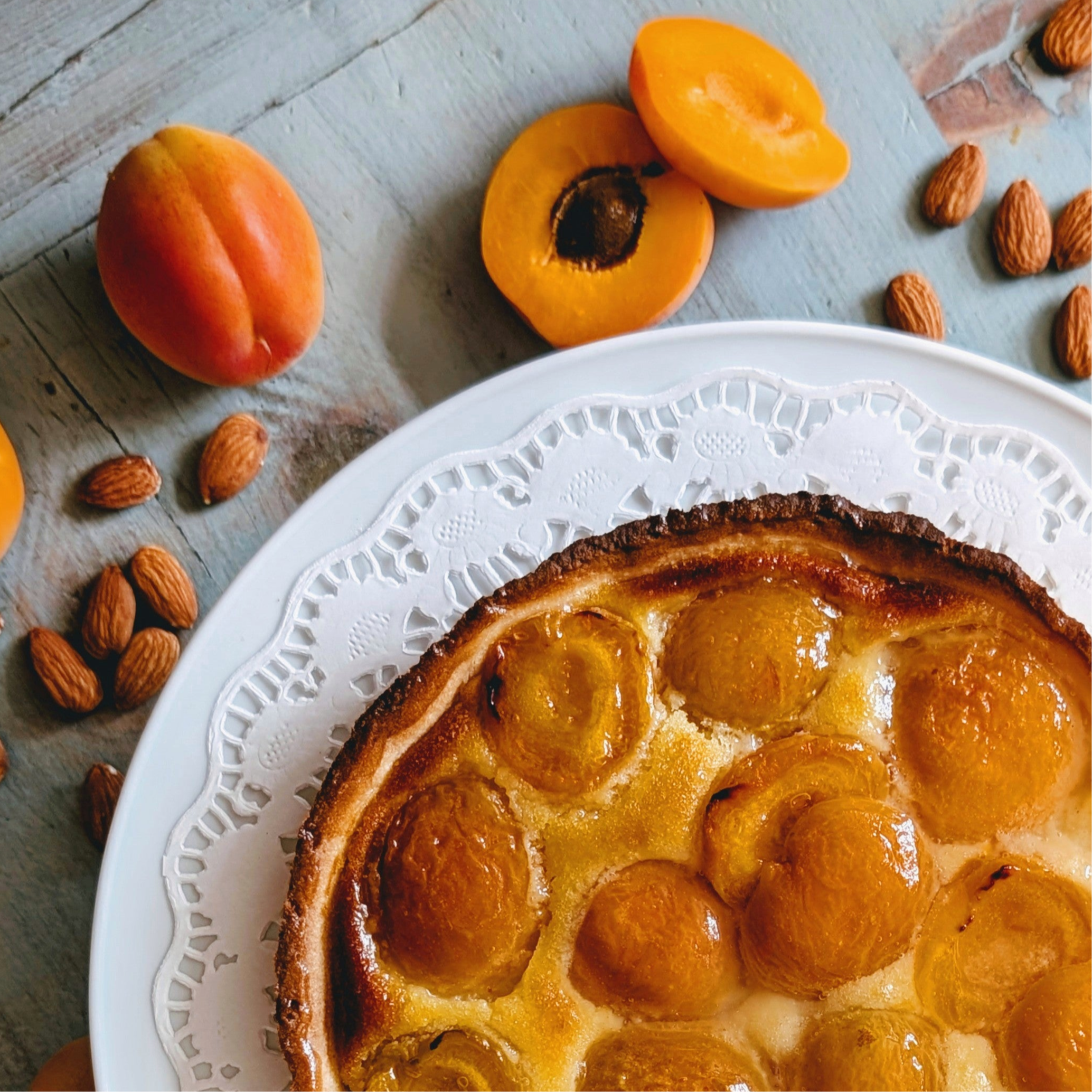 Apricot tart on a white doily plate with whole apricots and scattered almonds on a wooden table.