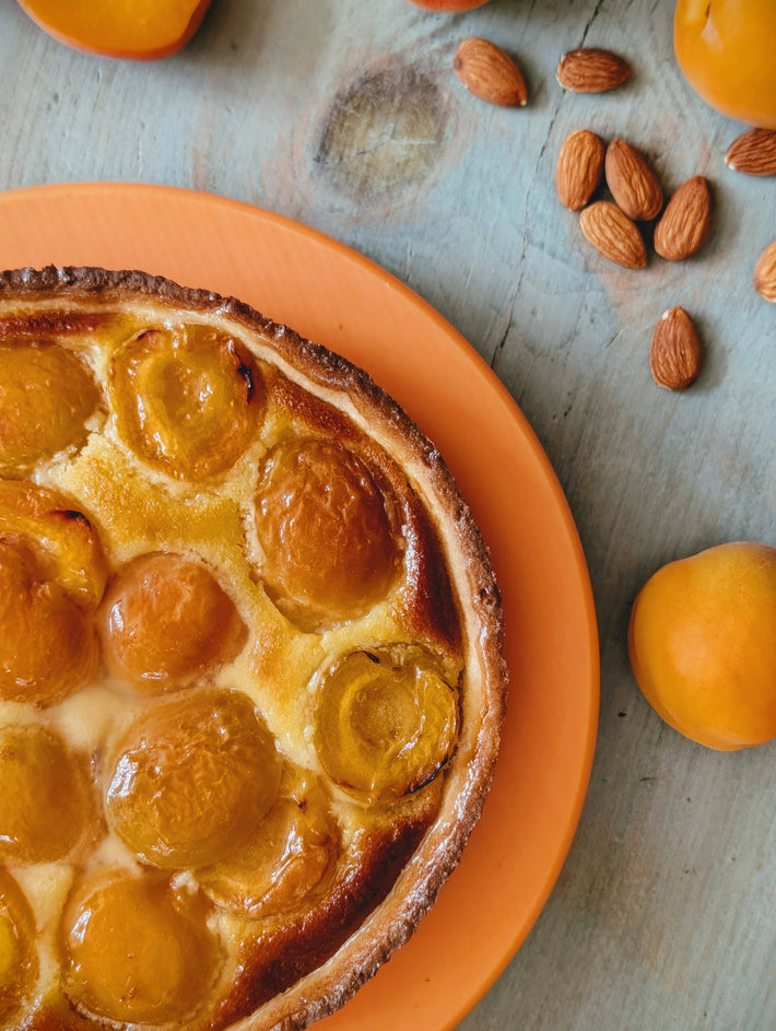 Apricot tart with glazed halves on an orange plate, almonds and whole apricots on a weathered wooden table.