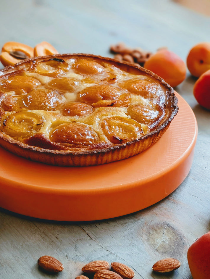 Apricot tart with glossy baked halves on an orange stand, whole apricots and almonds on wooden table