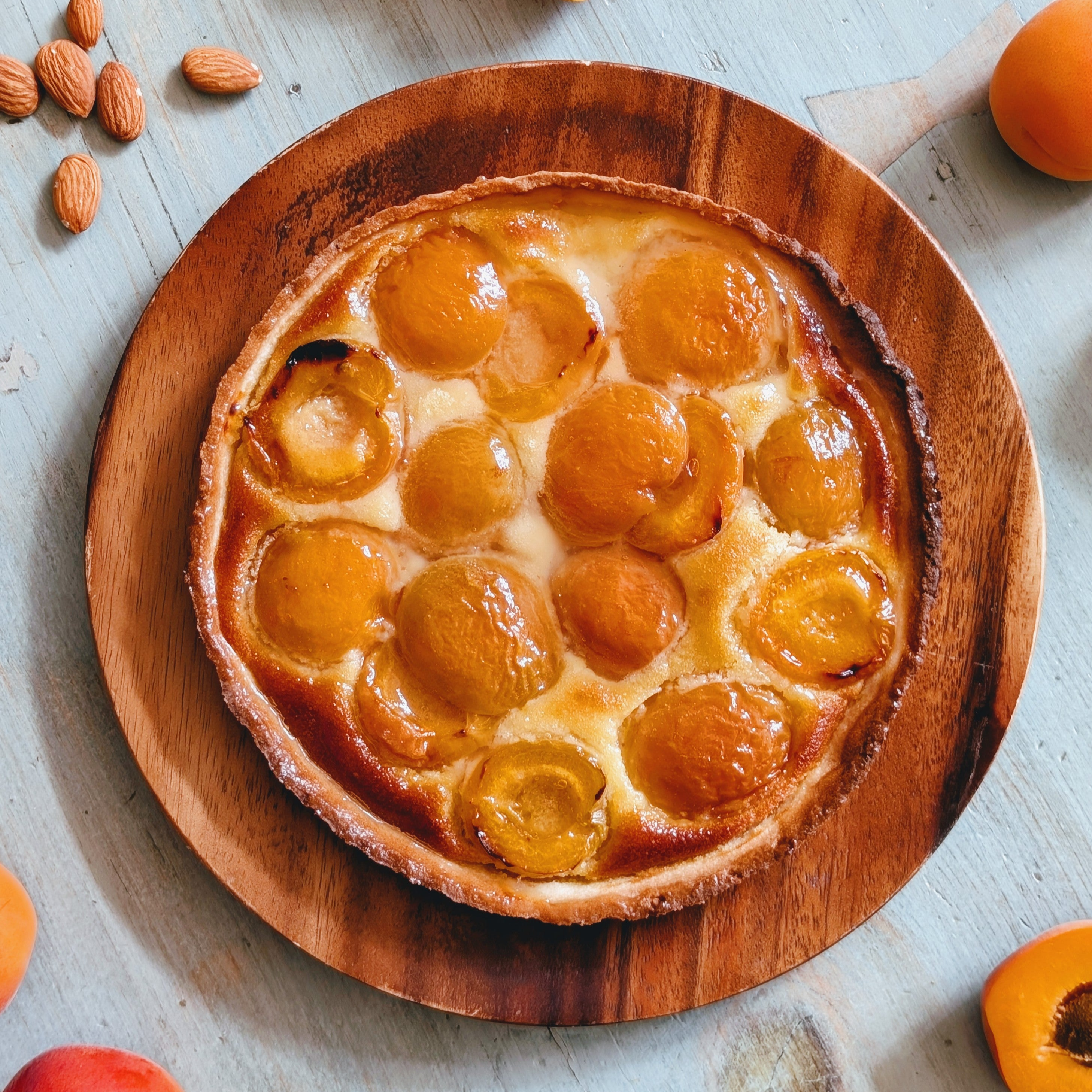 Apricot tart with glazed apricot halves on a wooden plate, whole apricots and scattered almonds nearby