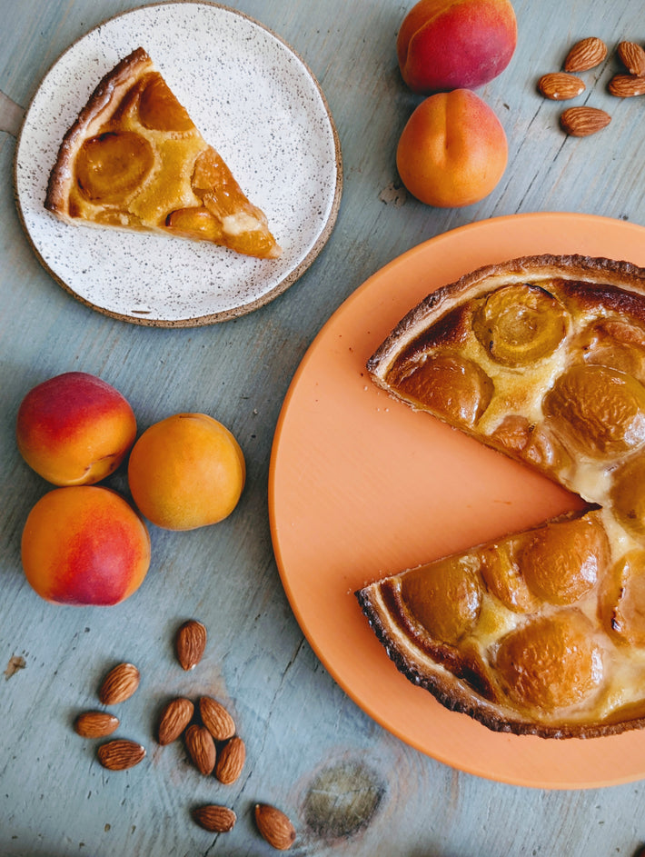 Apricot tart with two slices on orange and speckled plates, fresh apricots and scattered almonds on blue-gray wood.