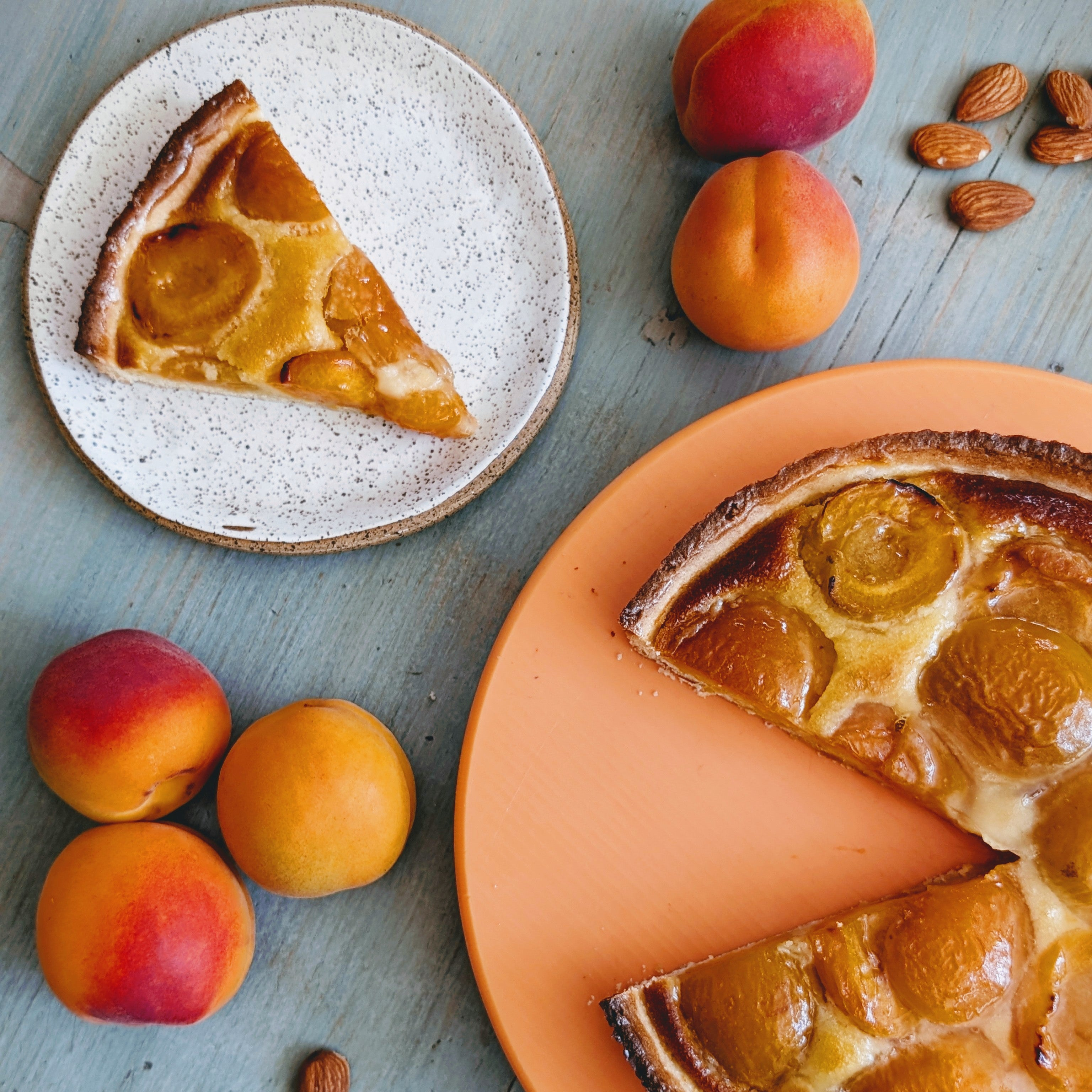 Apricot tart with two slices on plates, a whole tart, fresh apricots and almonds on a blue wooden table.