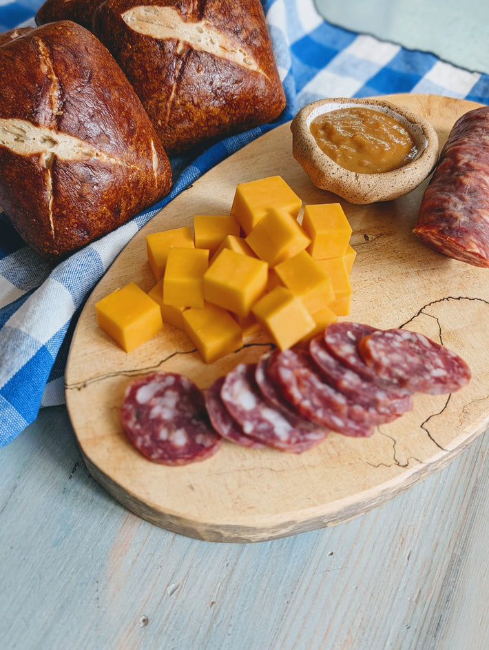 A wooden board displays cubes of cheese, sliced salami, a small bowl of spread, and fresh bread rolls, set on a checkered cloth.