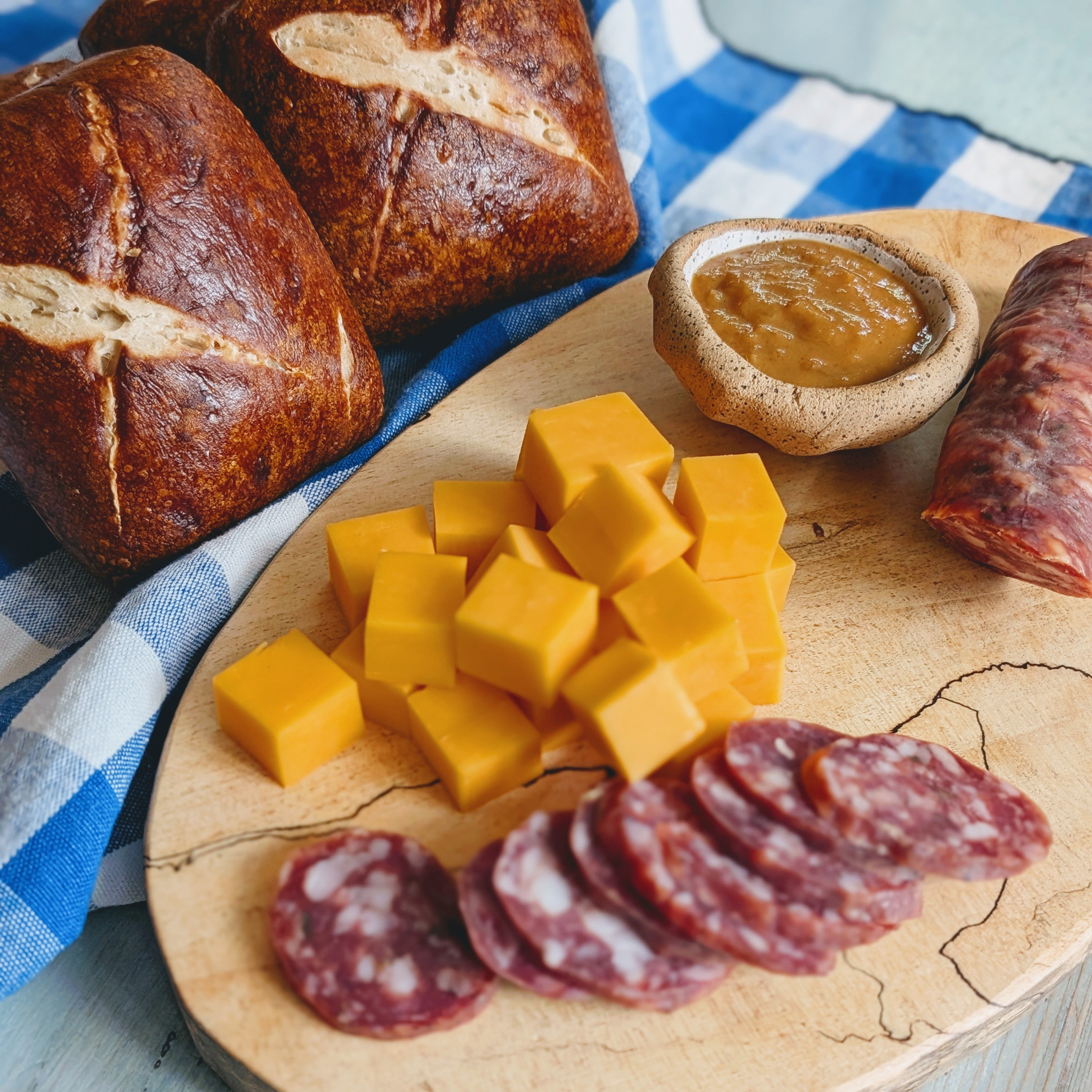 Cheese and charcuterie on a wooden board: cheddar cubes, sliced salami, mustard in a small bowl, and two rustic rolls on blue-check cloth.