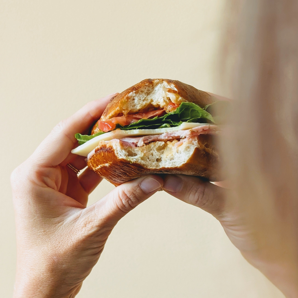 Hands holding a half-eaten sandwich with ham, cheese, lettuce, tomato on crusty bread.