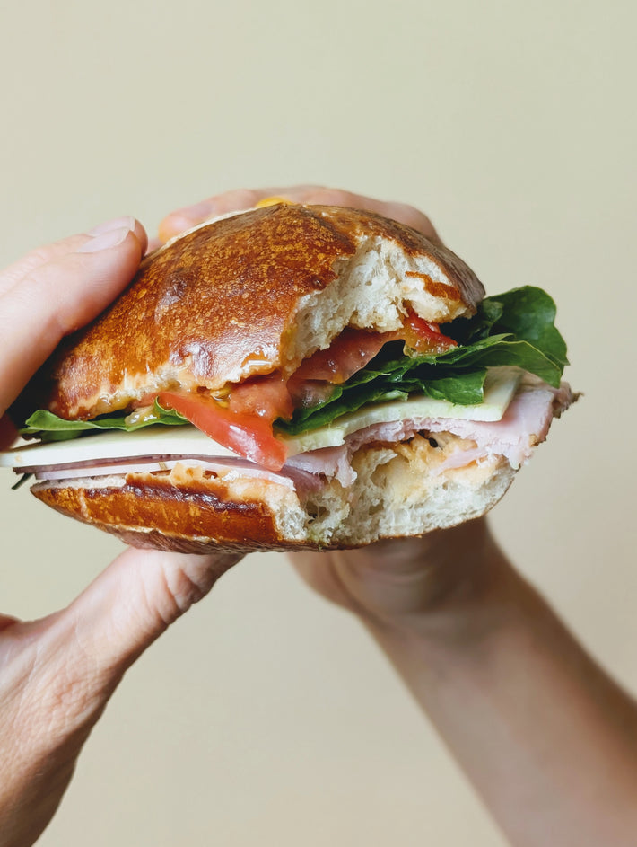 A hand holding a Bavarian pretzel sandwich with lettuce, tomato, and ham, featuring a golden-brown pretzel bun.