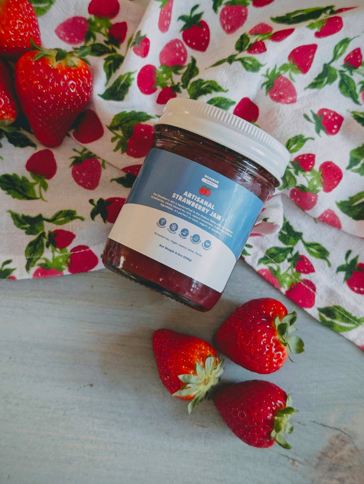 Glass jar labeled 'Artisanal Strawberry Jam' on strawberry-patterned cloth with three fresh strawberries on a light wood surface