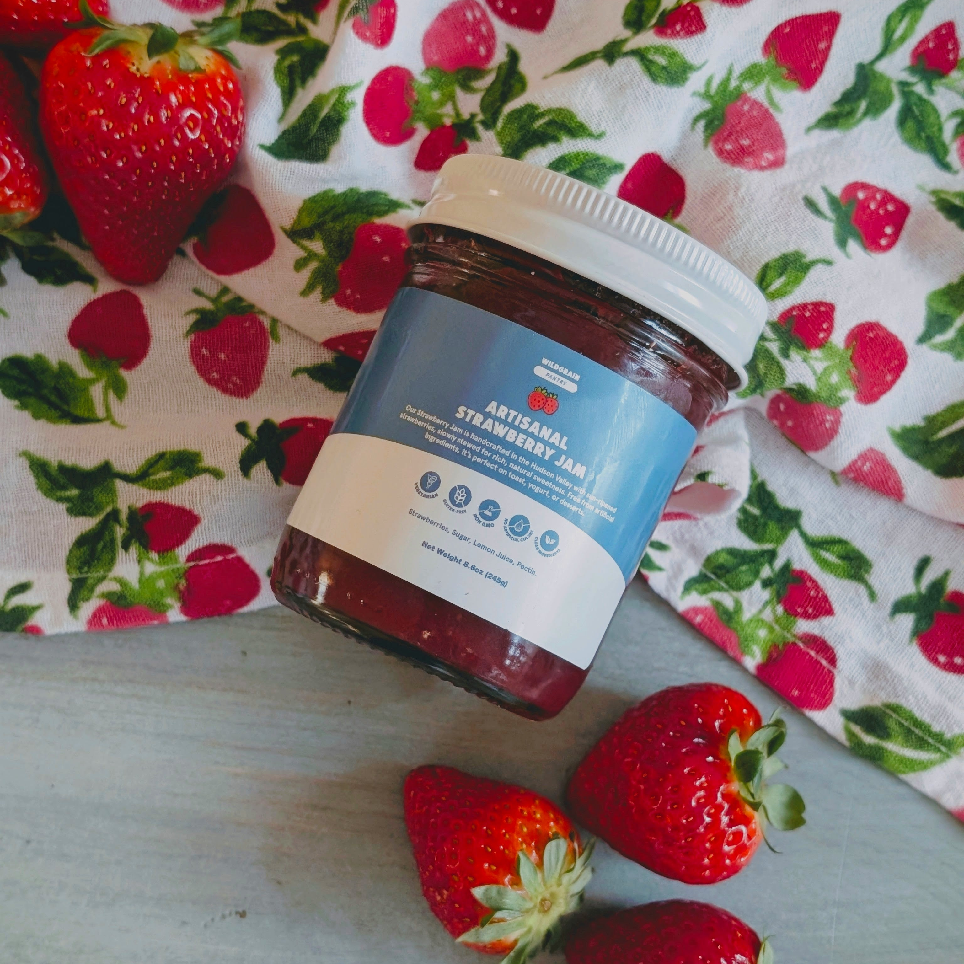 Glass jar labeled ARTISANAL STRAWBERRY JAM on strawberry-patterned cloth with fresh strawberries