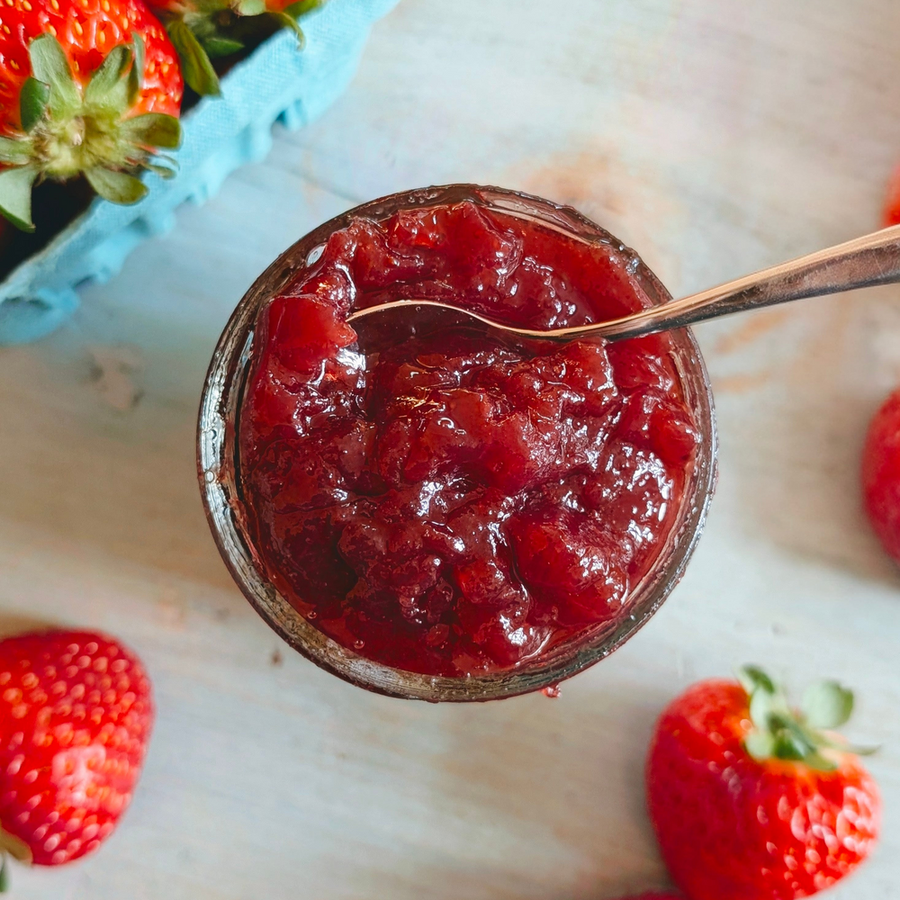Jar of strawberry jam with a spoon, surrounded by fresh strawberries on a light background