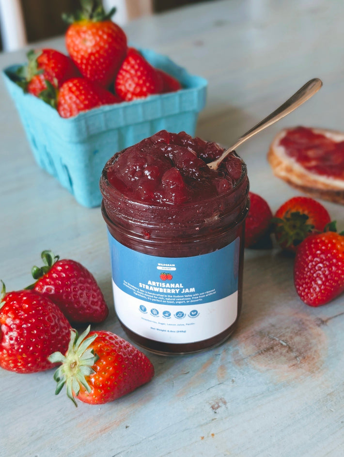 Jar of WILDGRAIN PANTRY ARTISANAL STRAWBERRY JAM with spoon, surrounded by fresh strawberries and a blue berry carton.