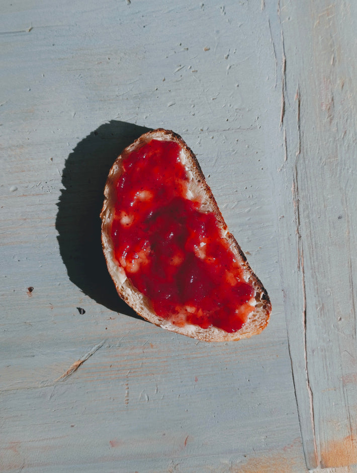 Crusty slice of bread spread with bright red fruit jam on a pale blue wooden board, casting a dark side shadow.