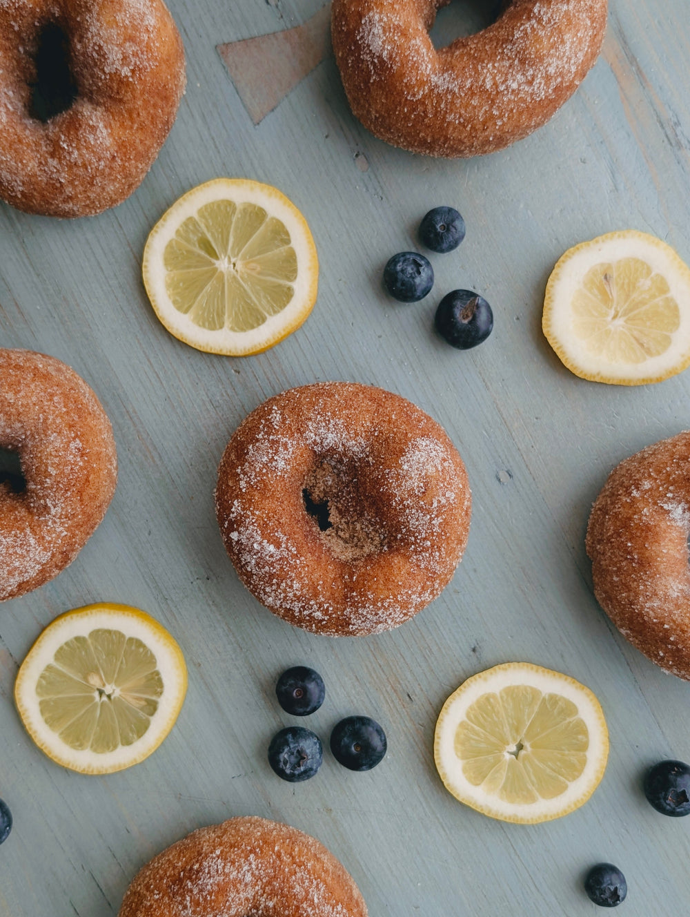 Top-down view of six blueberry lemon donuts with sugar coating, lemon slices, and fresh blueberries on a light blue wooden board