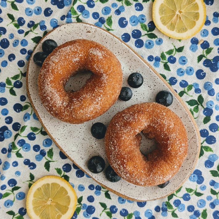 Top view of two Blueberry-Lemon donuts with sugar coating and fresh blueberries on a speckled plate over a blue floral cloth
