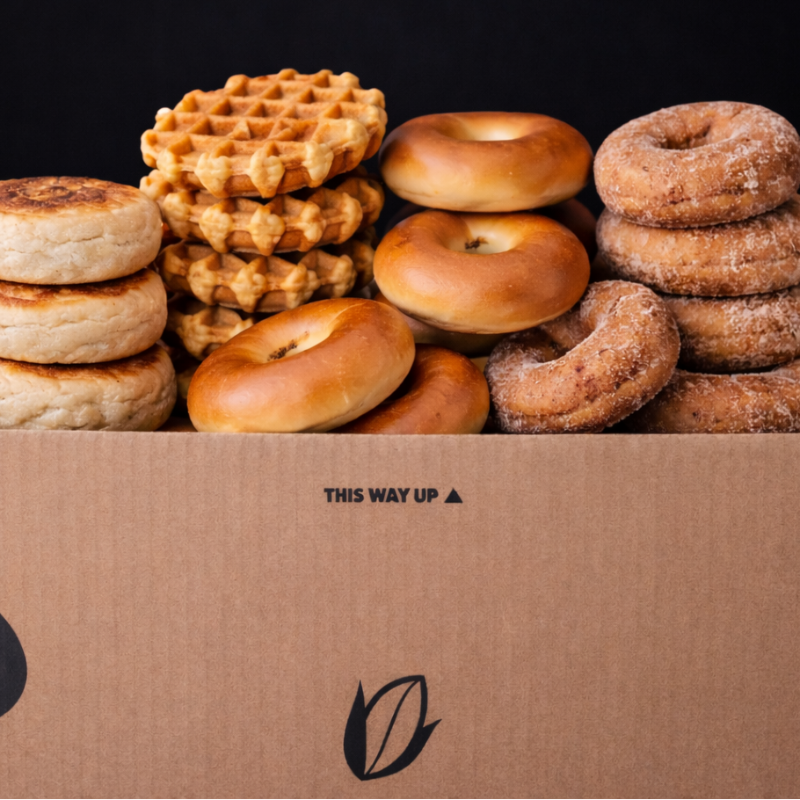Assorted pastries including waffles, bagels, and donuts in a cardboard box with 'This Way Up' label.
