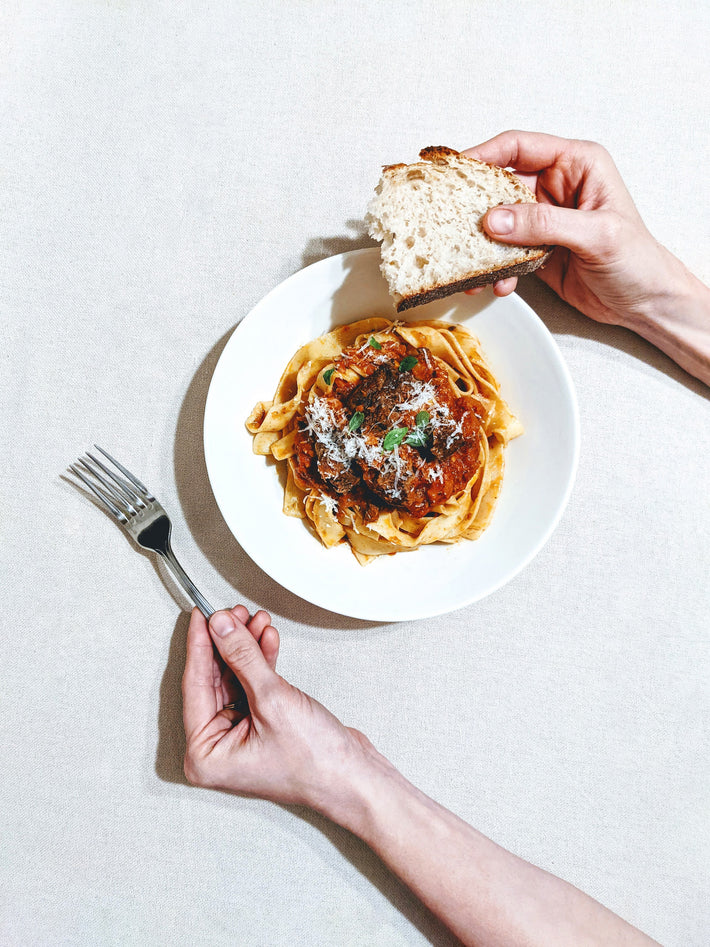 A person holding a piece of bread over a bowl of pasta with sauce and cheese, ready to eat.