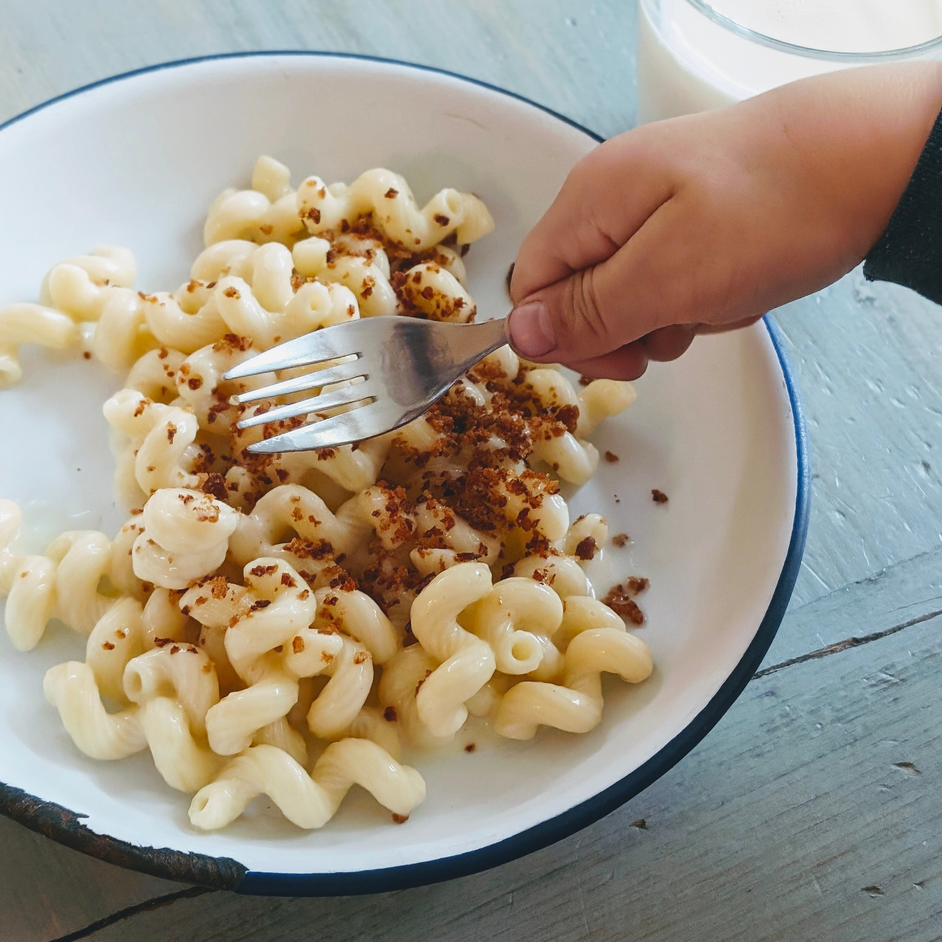 Child's hand holding a fork over a bowl of creamy spiral pasta topped with browned breadcrumbs