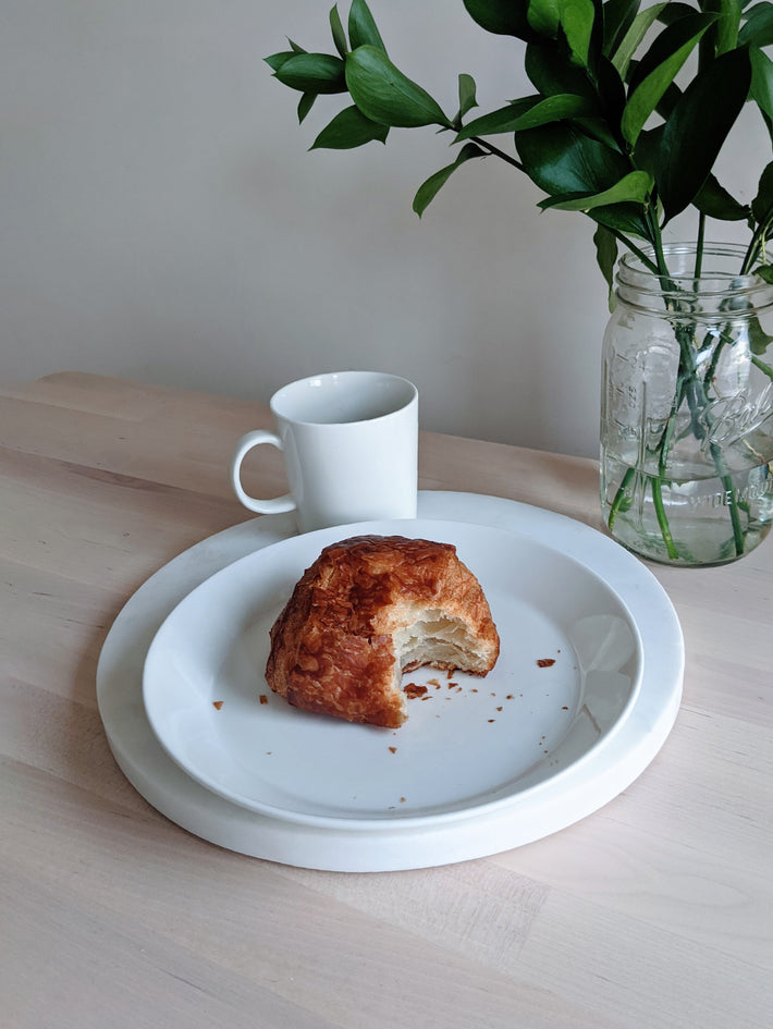 Partially eaten chocolate croissant with crumbs on a white plate, white mug behind it and a mason jar of greens.