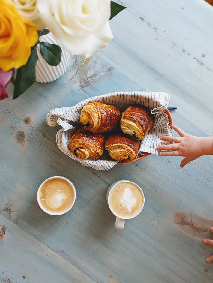 A basket of chocolate croissants with two cups of coffee on a table, surrounded by flowers.
