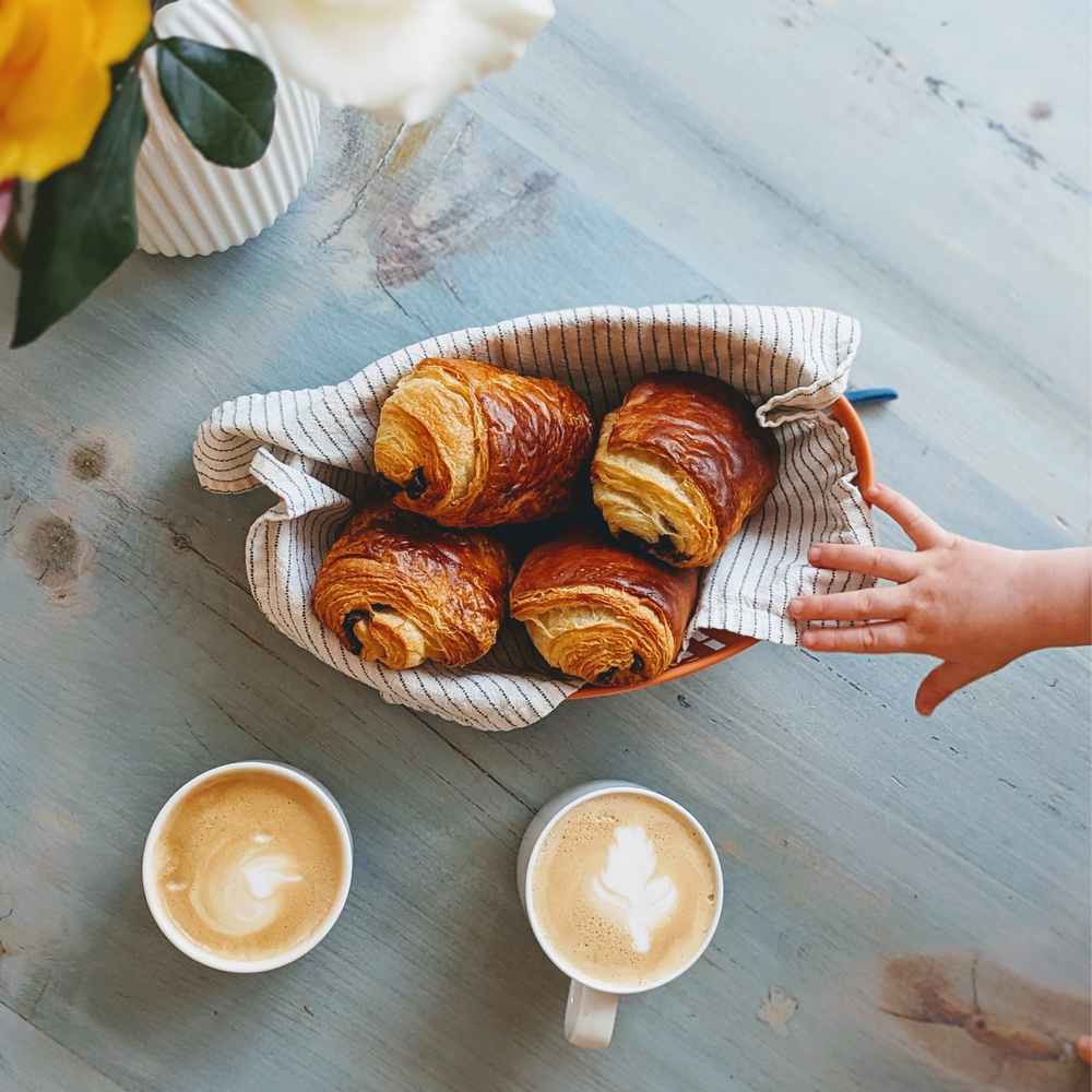 Basket of four chocolate croissants on striped cloth, two lattes with foam art, child's hand reaching from right on table