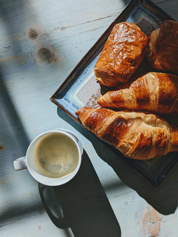 Overhead view of chocolate croissants on a rectangular plate showcasing flaky golden layers beside a white coffee cup on sunlit wood