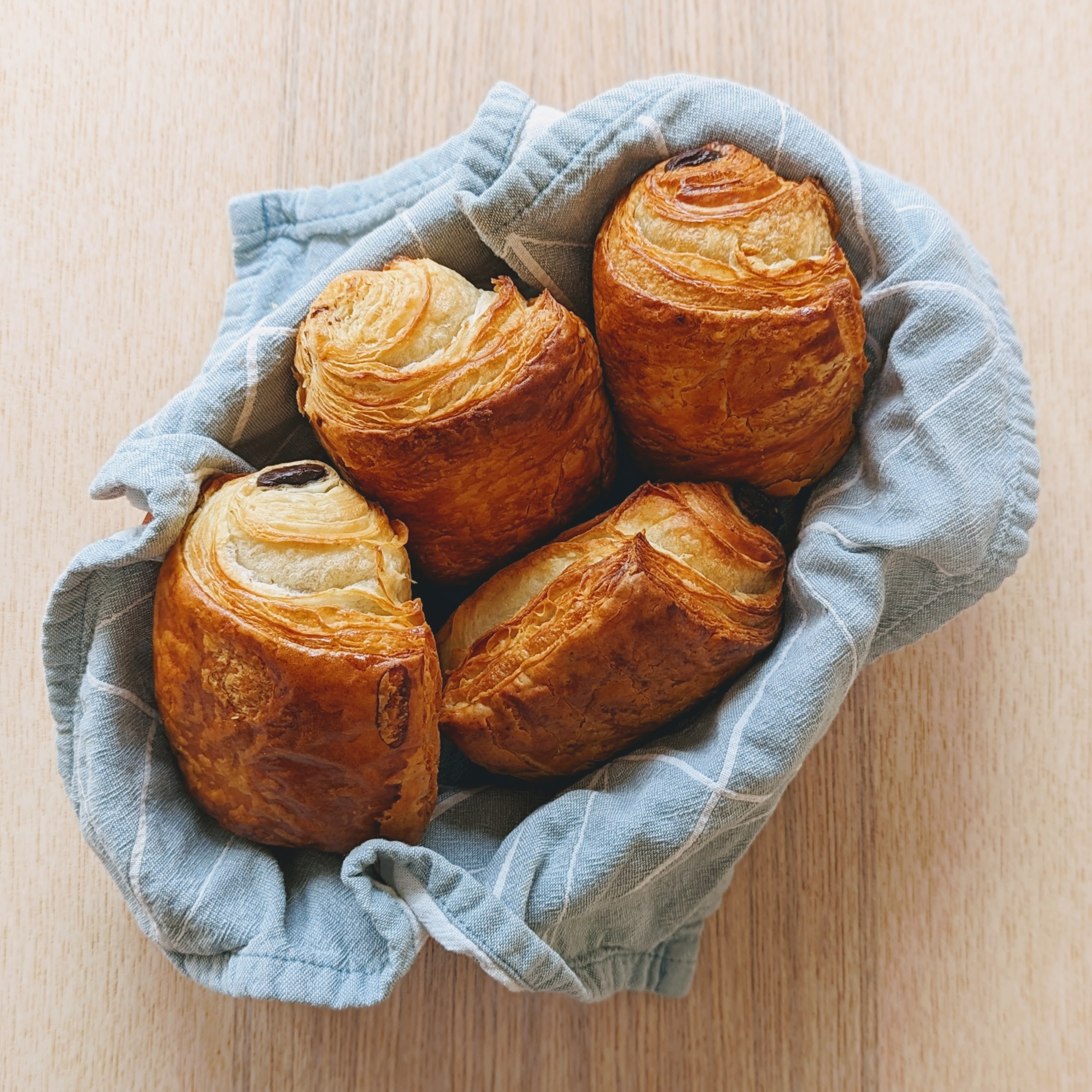 Four golden pain au chocolat nestled in a blue linen-lined basket on a light wood table.