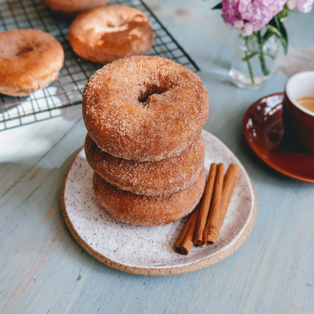 Stack of three cinnamon-sugar donuts on a speckled plate with two cinnamon sticks; coffee cup and cooling rack in background.