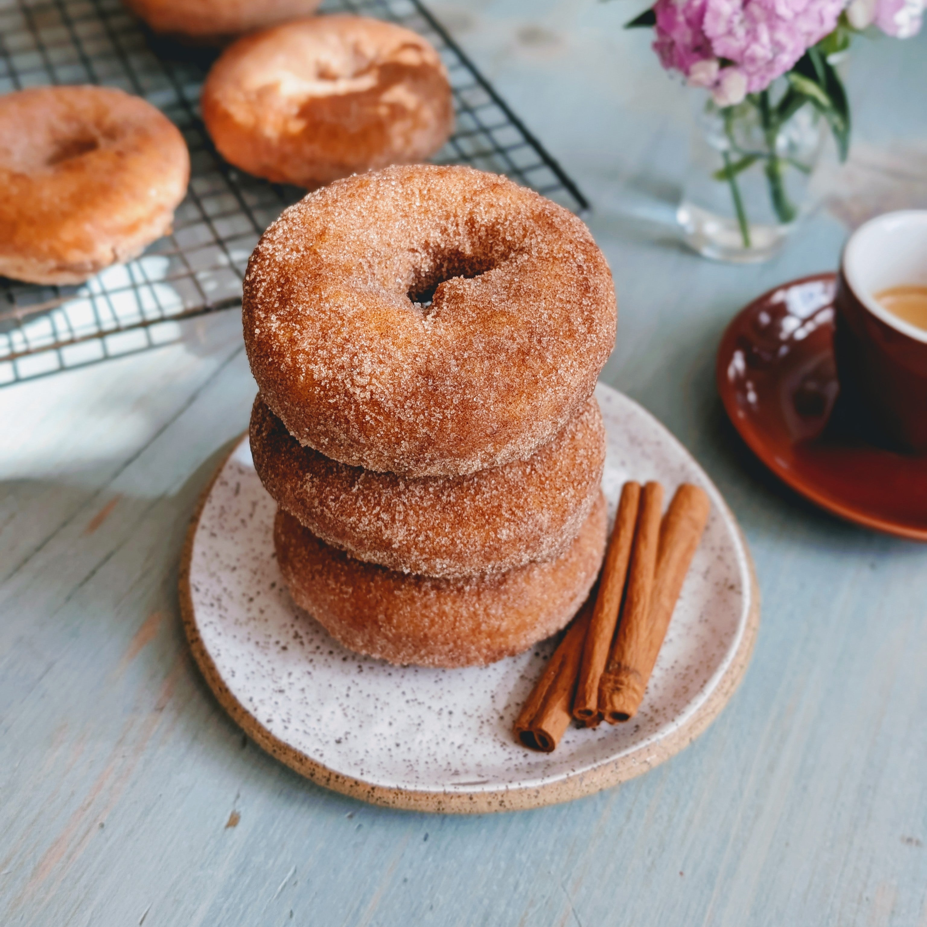 Stack of three cinnamon-sugar donuts on a speckled plate with two cinnamon sticks; coffee cup and cooling rack in background.