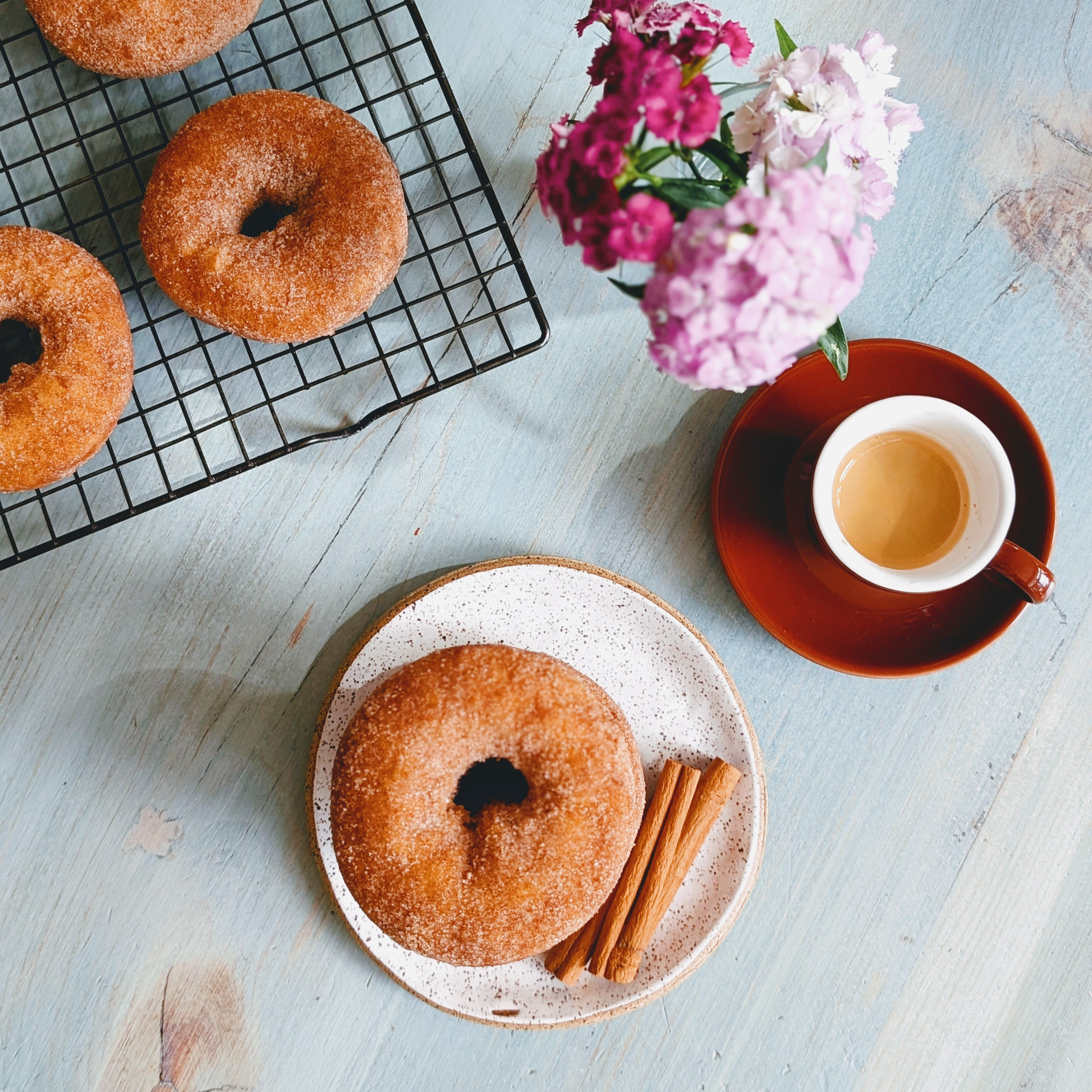 Sugared cinnamon donut on speckled plate with cinnamon sticks; cooling rack with donuts, espresso cup and pink flowers