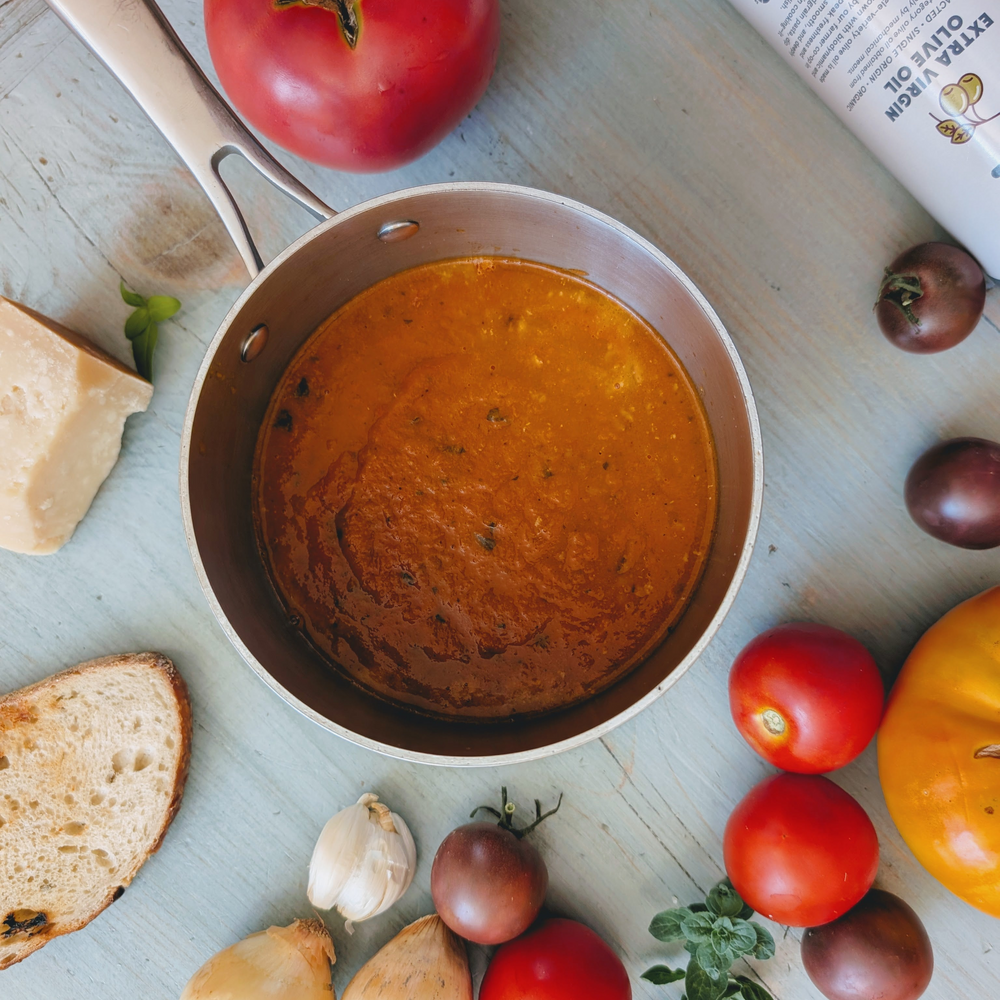 Saucepan of tomato soup on wood, surrounded by tomatoes, garlic, bread and cheese; tube labeled EXTRA VIRGIN OLIVE OIL