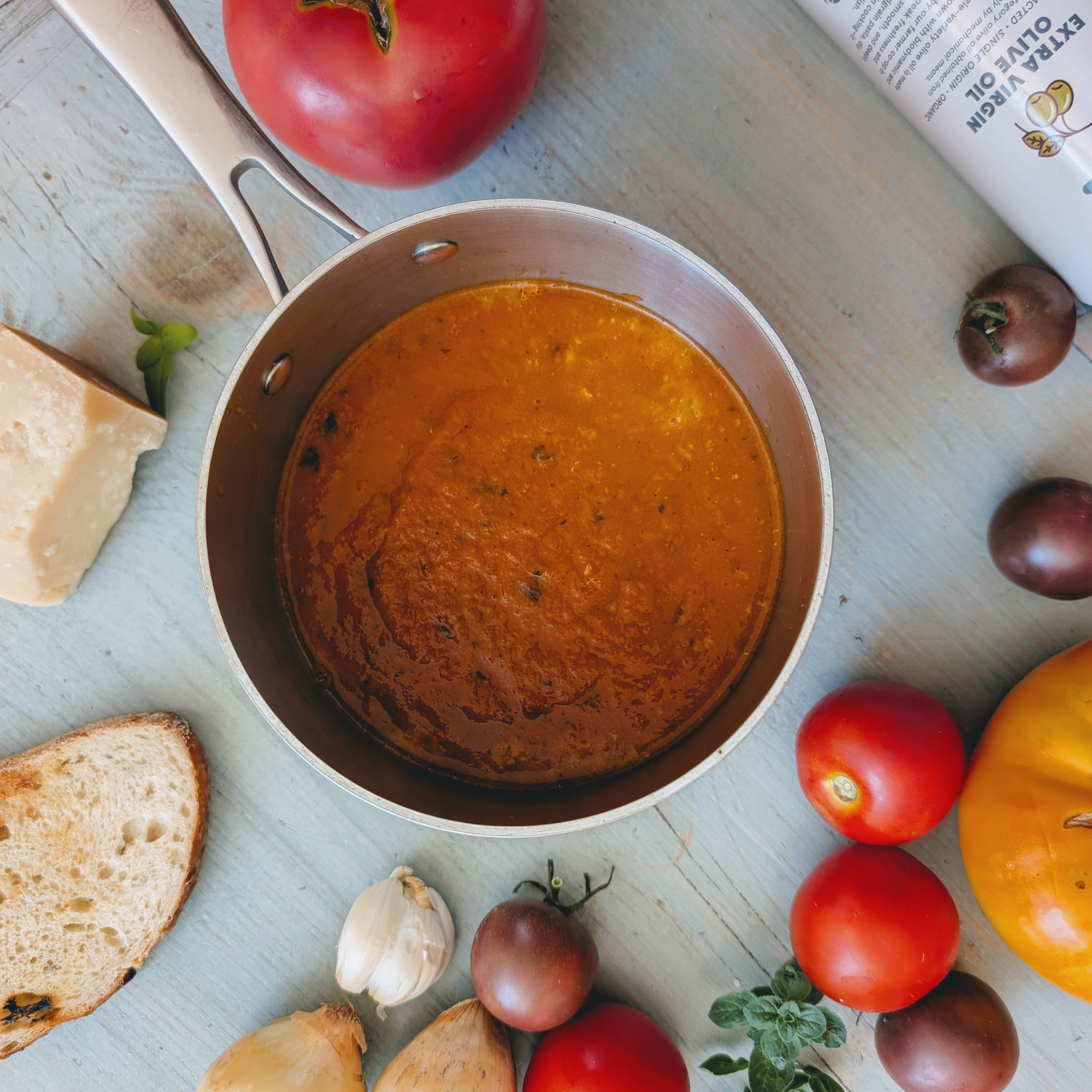 Saucepan of tomato soup on wood, surrounded by tomatoes, garlic, bread and cheese; tube labeled EXTRA VIRGIN OLIVE OIL