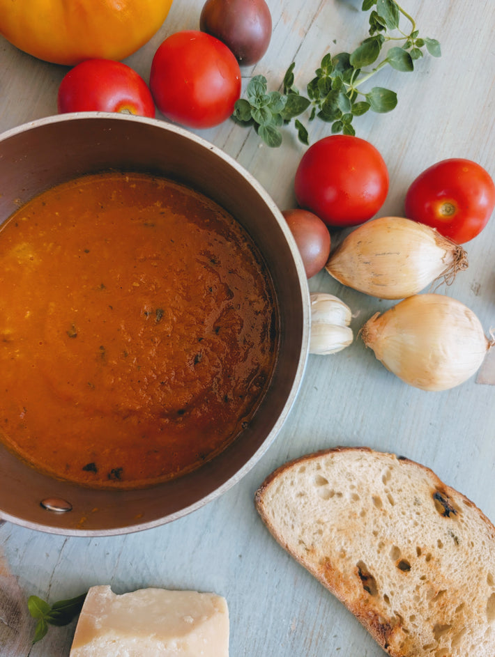 Pot of tomato soup with tomatoes, onions, garlic, herbs, toasted bread slice and cheese on a wooden table.