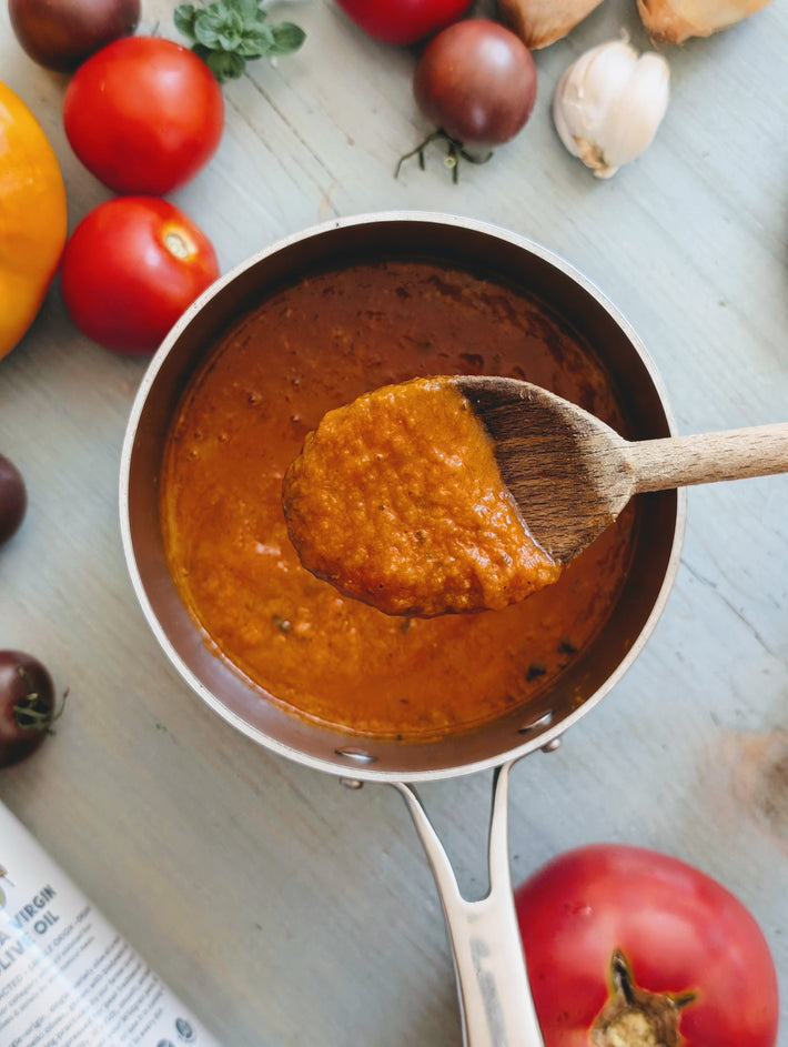 Pan of tomato sauce, wooden spoon holding a scoop; tomatoes, garlic, and a bottle labeled VIRGIN OLIVE OIL nearby.