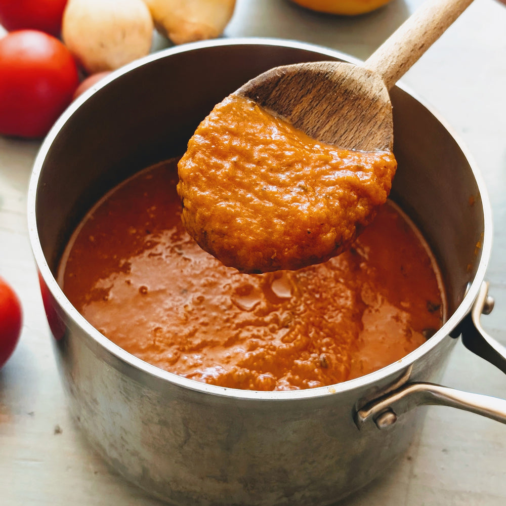 Saucepan of chunky tomato sauce being stirred with a wooden spoon, tomatoes and onions visible in background.